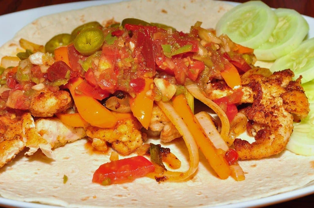 An open flour tortilla topped with spiced chicken, strips of red and yellow pepper, onion and sliced jalapeño