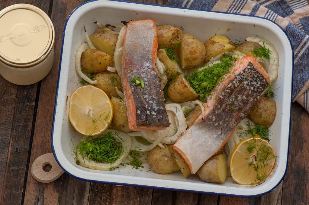 Two skin-on salmon fillets on a bed of roasted baby potatoes and onions with lemon halves in an enamel baking dish